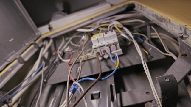 Exposed electrical wires tangle around vintage circuit breakers inside a dusty metal distribution box. Camera pulls back to reveal chaotic cabling and potential safety hazards.