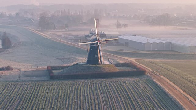 Aerial view of a windmill standing tall amidst agricultural fields and farm buildings, casting long shadows in the early morning light, Beesel, Limburg, Netherlands.