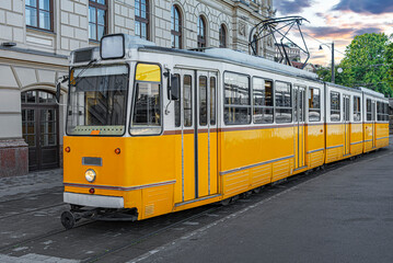 Naklejka premium A classic yellow and white electric tram sits on tracks alongside an ornate historical building under a dramatic sunset sky.