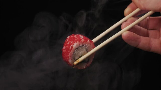 Male hand holds California sushi roll with red roe using wooden chopsticks. Thick white smoke swirls around Japanese appetizer creating dramatic presentation effect.