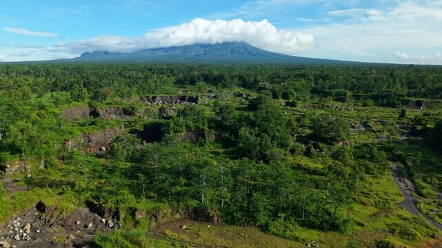 Aerial view of the Woro River weaving through a landscape of lush green trees, fields and a majestic merapi mountain under a partly cloudy sky, Klaten Regency, Indonesia.
