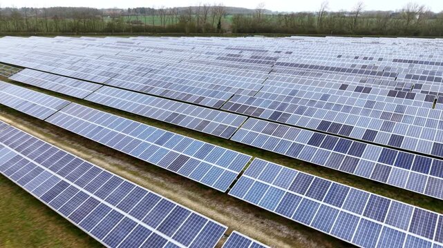 Aerial view of a vast solar farm displaying a grid of dark blue panels contrasting with the light green and brown grassy land, former RAF Alconbury, United Kingdom.