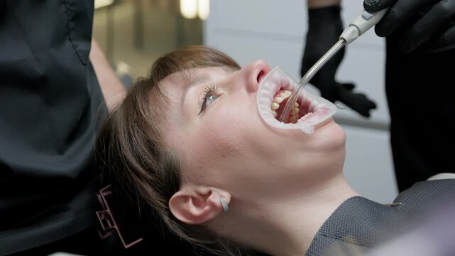 Professional dentist performing a dental procedure on a female patient with a cheek retractor, using a saliva ejector and other specialized tools inside a modern and clean clinic office