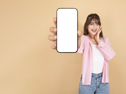 Excited young woman showing smartphone with blank screen toward camera, wearing pink shirt and denim, standing against beige studio background with wide copy space. Mobile app mockup concept.