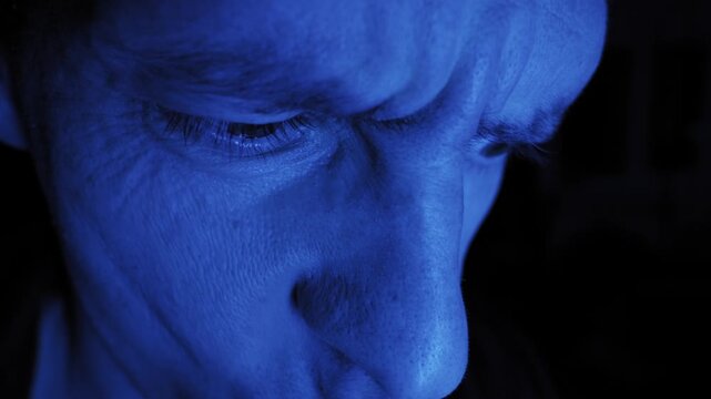 Caucasian male face looks downwards showing signs of stress and fatigue. Strong monochrome blue light highlights skin texture with deep dramatic shadows.