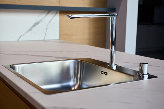 High-end installation with stainless steel sink installed in white marble look surface. Rectangular single bowl brushed finish sleek, L shaped chrome faucet and deck mounted soap dispenser.