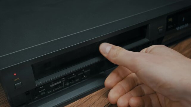 A male hand inserts a black VHS cassette into the slot of a vintage VCR player on a wooden table. Concept of watching old movies, nostalgia, and analog technology history.
