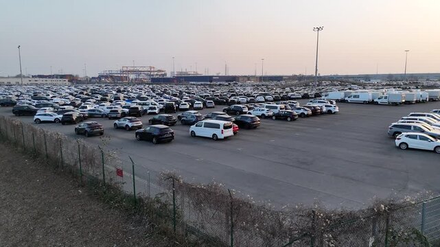 Drone crosses the fence and reveals the full extent of the vehicle terminal with cranes and industrial backdrop.