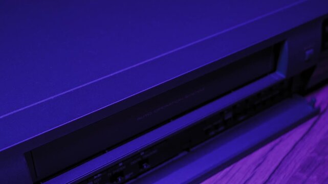 Male hand inserts black VHS cassette into vintage video recorder slot. Dark room illuminated by vibrant blue and purple neon lighting creates nostalgic 80s atmosphere.