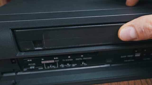 Male hand inserts black VHS cassette tape into vintage VCR player slot. Finger presses play button on retro electronic device with auto operation system label.
