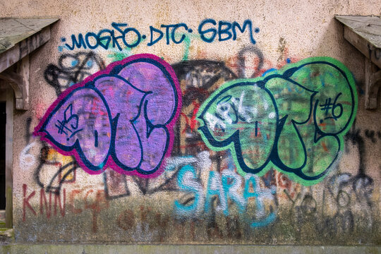Urban graffiti adorns a weathered concrete wall. Bold purple and green tags dominate the surface. Faded spray paint streaks add texture to the scene. Weathered brickwork frames the graffiti display.