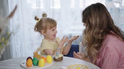Obraz premium Bright kitchen with soft light filtering through curtains. Young girl smiles, hands raised in joy. Mother sits beside her, sharing a sweet moment. Easter eggs, small cake. Ideal for family, holiday