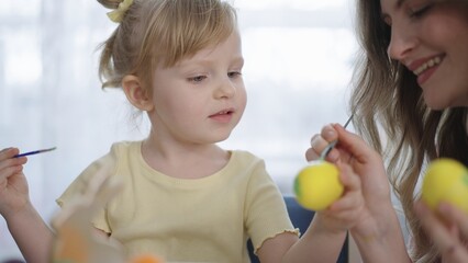 Fototapeta premium Little girl paints eggs with mother's help. Both smile warmly during joyful activity. Soft lighting enhances cozy, playful atmosphere. Ideal for parenting, holiday, or family lifestyle scenes