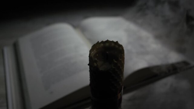 Close-up of a hand lighting a honeycomb beeswax candle with a match. The flame illuminates an out-of-focus book in a dark room, creating a studious or mystical atmosphere.