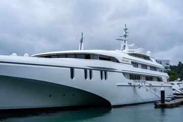 White luxury yacht floats calmly in harbor waters. Cloudy sky looms overhead, softening the scene. Modern design features sleek lines and large windows. Water reflects yacht's clean shape.