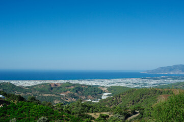 Obraz premium horizontal shot overlooking a vast valley filled with white greenhouse complexes stretching towards the deep blue sea, bordered by green forested hills and distant mountain ranges under a clear sky.