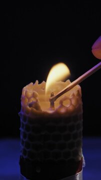 A hand lights the wick of a honeycomb beeswax candle using a burning match against a dark background. Concept of warmth, spiritual rituals, relaxation, and natural eco-friendly home decor.
