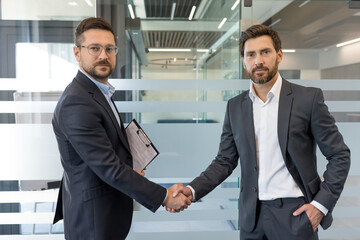 Two confident businessmen in formal wear are shaking hands, symbolizing agreement, partnership, and successful collaboration during a business meeting in a modern office environment