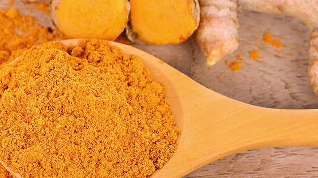 Turmeric powder in wooden spoon with sliced turmeric root from top view, organic spice used in cooking and traditional medicine.