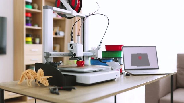 A 3D printer is actively creating a small red object on its build plate, with spools of filament visible in the background