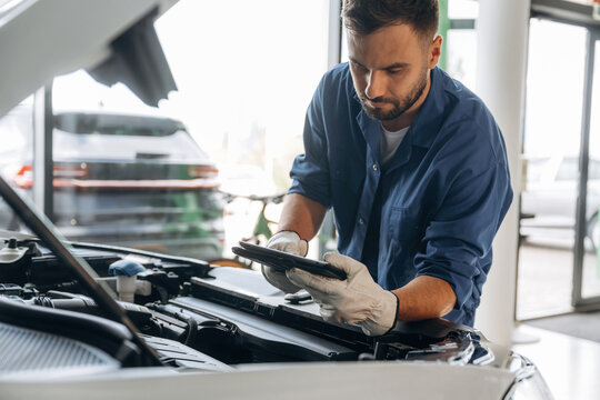 Black digital tablet, hood is opened, fixing the automobile. Male mechanic is working in the modern car workshop