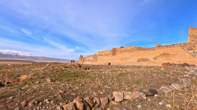 Wide view of Ani city walls in Kars, Turkey. Medieval fortifications stretch across vast steppe landscape with mountains in background under clear blue sky, highlighting history and architecture.