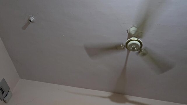 A spinning white ceiling fan from below. Its three blades are blurred by motion, and a clear shadow is cast against the off-white ceiling. An empty light socket is visible nearby on the left.