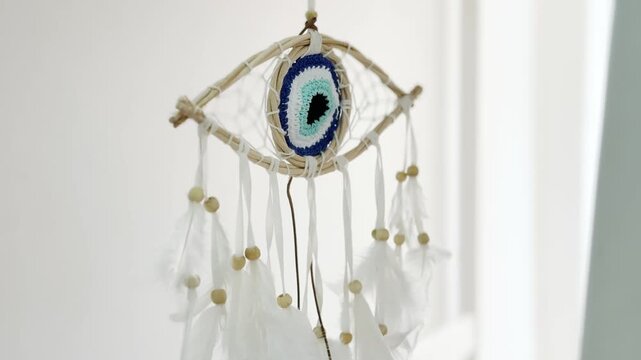 Beautiful handmade dream catcher shaped like a nazar eye with white feathers and wooden beads gently swaying in the sunlight against a white wall for spiritual protection and good luck