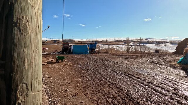 Muddy farmyard with wheelbarrow, farming equipment and open countryside. Snow patches in the background under clear blue sky, showing rural work environment and everyday agricultural life.