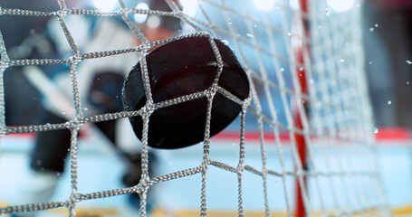 Close-up action shot of an ice hockey puck hitting the goal net during a scoring moment. Dynamic sports scene with ice spray and a blurred hockey player in the background. © Jag_cz