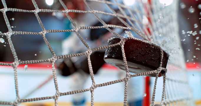Close-up action shot of an ice hockey puck hitting the goal net during a scoring moment. Dynamic sports scene with ice spray and a blurred hockey player in the background.