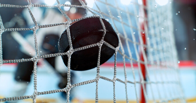 Close-up action shot of an ice hockey puck hitting the goal net during a scoring moment. Dynamic sports scene with ice spray and a blurred hockey player in the background.