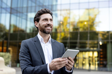 Fototapeta premium Confident businessman with a beard smiling, holding a digital tablet while thinking about future success and innovative business strategies in front of a modern building