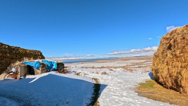 Snowy path in rural area leading between old structures and haystack toward a lake. Clear blue sky and winter light create a peaceful countryside scene with depth and natural composition.