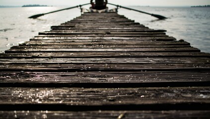 Obraz premium Leading wooden pier stretching to rowboat at lakeshore with person wearing dark coat, oars glinting