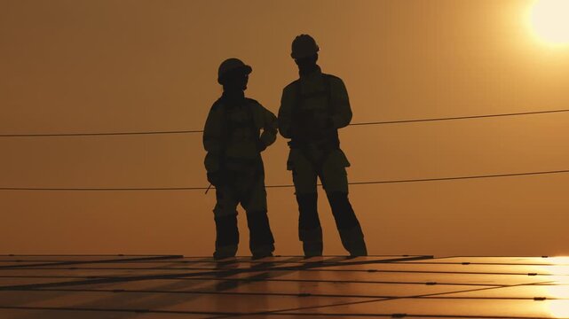 Two solar technicians sitting on rooftop photovoltaic panels at sunset discussing inspection results and pointing toward the solar installation area.