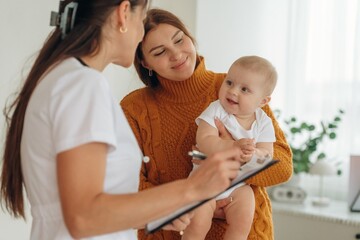 Using pen, writing information. Mother with her little baby daughter is in clinic with doctor at appointment
