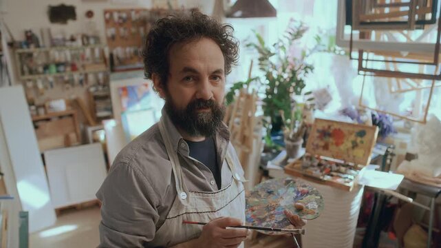 Portrait of male painter in apron holding palette and brush, looking at camera while standing in his creative studio, surrounded by artworks, tools and frames
