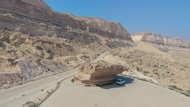 Aerial view of a white SUV parked under a large mushroom-shaped rock formation beside a winding mountain road near Salalah, Oman.