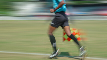 Fast linesman chasing play along sideline with chequered flag capturing urgency focus speed judgment on grassy pitch during football game