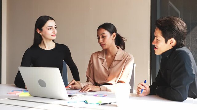 Three business professionals discussing charts and reports while analyzing information on laptop during collaborative office meeting.