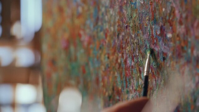Extreme close up view of painters hand using fine tip brush to apply oil paint to colorful textured canvas, creating abstract impressionist artwork in a sunlit studio