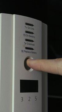 Finger presses power button on a white uninterruptible power supply causing the light to activate. Vertical. Concept of electrical safety, power outage preparation, and hardware maintenance.