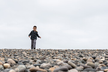 Obraz premium Little boy walking on a rocky beach under cloudy sky with copy space