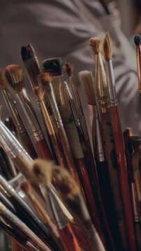Collection of used professional paintbrushes with different bristle types and wooden handles gathered in container in creative studio, artist working in background. Vertical close up shot