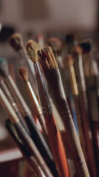Variety of used paintbrushes with different bristle types and sizes, organized in holder in softly lit art studio. Vertical clip, close up view