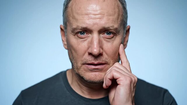 A middle-aged man with a serious expression touches his face, appearing contemplative or concerned. He has short, grayish hair, stubble, and wears a dark shirt against a light blue background.