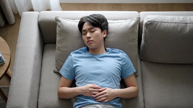 A young person sleeps on a beige sofa with hands clasped on stomach, wearing a blue T-shirt, in a cozy room with curtain, side table, and book. Image sequence shows peaceful rest.