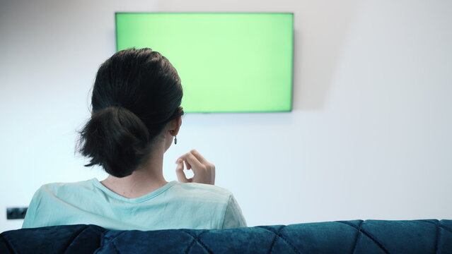 Girl Relaxing on the sofa, Sits in Font of a Geen Sreen TV, Eats Chips and Switches Channels, Attentively Watches Information on TV, Green Chroma Key Screen TV, have fun and enjoy