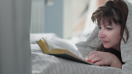 Naklejka premium woman lies on the bed and reads a book, covered with a blanket, have fun and enjoy